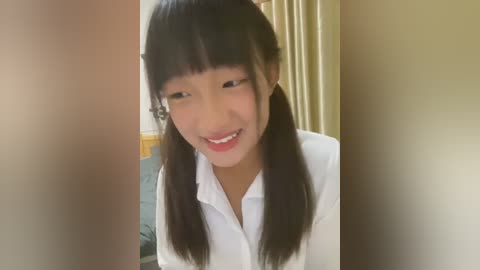 Media: Video of a young East Asian girl with straight black hair in pigtails, smiling broadly, wearing a white shirt, indoors, with beige curtains and a wooden object partially visible in the background.