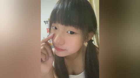 Media: Video of an Asian girl with straight black hair in pigtails, wearing a white shirt, using her index finger to touch her eye, indoors with blurred background.