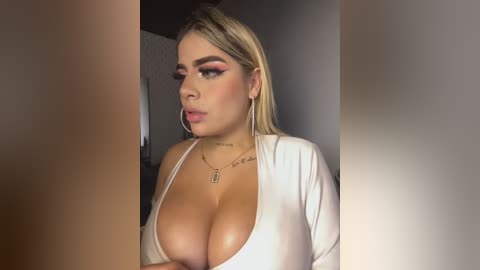 Media: Video of a curvy, light-skinned woman with blonde hair, wearing a plunging white top that reveals large breasts, and multiple tattoos on her chest.