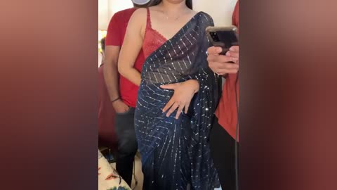 Media: Video of a woman in a red sleeveless top and dark blue saree with white sequins, taking a selfie in a crowded indoor setting.