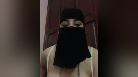 Media: Video of a woman with long black hair, wearing a black veil covering her face, a white spaghetti-strap top, and standing in front of a red door.