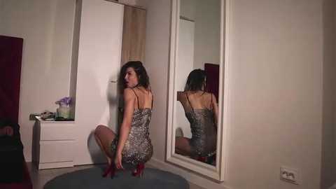 Media: Video of a woman in a short, glittery dress squatting in front of a mirror, her reflection visible, in a dimly lit bedroom with white walls, a dresser, and a purple flower arrangement.