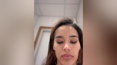 Media: Video of a young Asian woman with long, dark hair and light skin, wearing black eyeliner and mascara, looking downward with closed eyes, in a white-walled hallway with a wooden door and ceiling tiles.