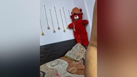 Media: Video of a red teddy bear with a yellow hat and a red scarf, leaning against a white wall adorned with brass candleholders, in a cozy, colorful room with a patterned rug.