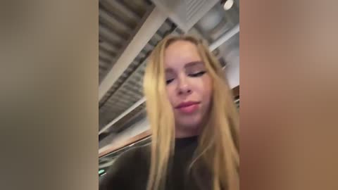 Media: Video of a young woman with long blonde hair, wearing a black top, making a pouty face in a modern, indoor setting with exposed metal beams and industrial ceiling lights.