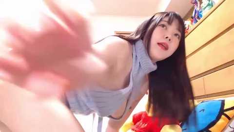 Media: Video of an East Asian woman with long black hair, wearing a light blue sleeveless top, leaning forward, capturing a playful, energetic moment in a brightly lit bedroom.