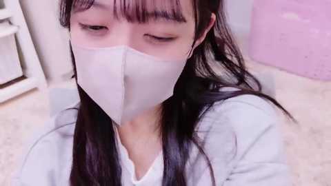 Media: A video of an East Asian woman with long black hair, wearing a white face mask and light blue jacket, sitting indoors with light-colored walls and a white shelf in the background.