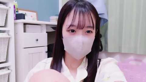 Media: Video of an Asian woman with long black hair, wearing a white mask and white blouse, holding a baby, in a light-colored room with a white desk and wicker baskets.