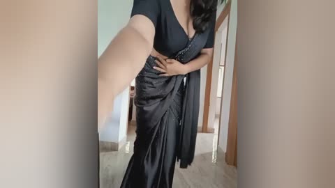 Media: Video of a South Asian woman with medium skin tone, wearing a black sari and blouse, standing in a room with wooden furniture and light-colored walls.