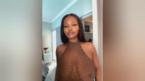 Media: Video of a confident Black woman with medium skin tone and long straight hair, wearing a sheer brown halter top that reveals her ample breasts, standing in a modern, well-lit bedroom with white walls, a bed, and a small table with red flowers.