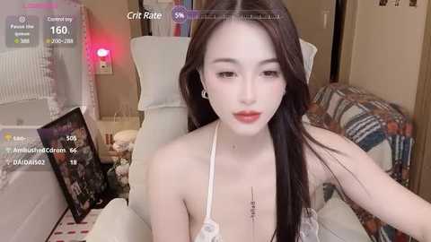 Media: Video of a young, fair-skinned, slender Asian woman with long black hair, wearing a white halter top, sitting on a white chair in a cozy room.