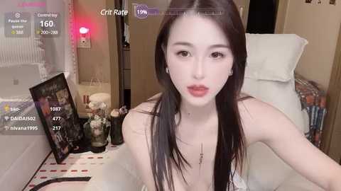 Media: Video of an East Asian woman with pale skin and long black hair, wearing a white top, sitting on a bed. Background includes a framed photo, a nightstand with a lamp, and a white pillow.