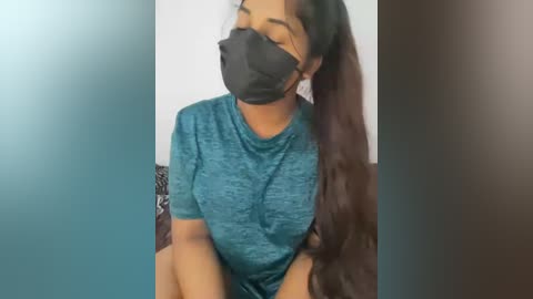 Media: Video of a woman with medium brown skin, wearing a black face mask, blue short-sleeve shirt, and ponytail, sitting indoors with a blurred background.