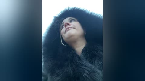 Media: Video of a woman with light skin, short dark hair, and wearing a black fur-trimmed coat, looking upward with a contemplative expression. Background is blurred, creating a dramatic, intimate atmosphere.