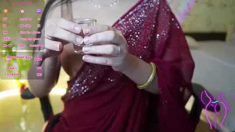 Media: Video of a woman in a deep red saree with sequins, holding a glass of water. The background is blurred, showing a green plant and a wooden chair.