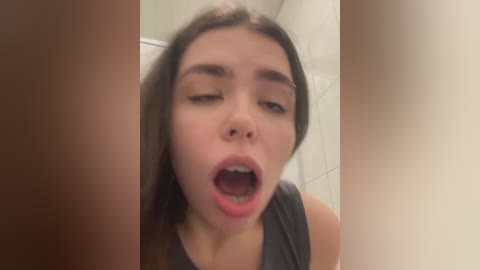 Media: A video of a young woman with long brown hair and light skin, wearing a gray tank top, looking shocked and mouth open in a bathroom.