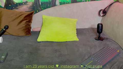 Media: Video of a minimalist room with a grey bedspread, a lime-green pillow, a brown pillow, a black microphone, and a black headset, with a green keyboard and a yellow remote control on the bed.