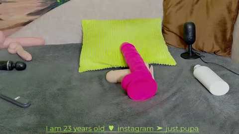 Media: A video shows a woman on a grey bed, wearing a bright pink dildo. Surrounding her are a yellow pillow, a black vibrator, a white massager, and a brown cushion.