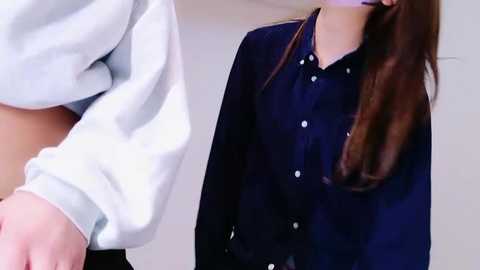 Media: Video of a woman in a navy blue button-up shirt, standing indoors. Her long brown hair cascades over her shoulders. She holds hands with a man in a white sweater, visible only from the waist down.