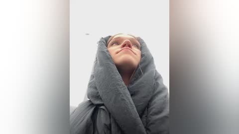 Media: Video of a young woman with light skin and short blonde hair, wearing a grey hoodie, looking upwards with a contemplative expression. The background is a white wall, creating a minimalist, serene atmosphere.