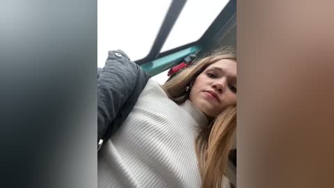 Media: A video shows a young woman with long, straight brown hair, wearing a white ribbed sweater, looking down from a car window. The background reveals a blurry, blue-tinted car interior.