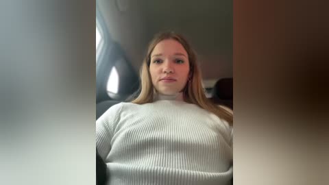 Media: A video of a young Caucasian woman with straight, shoulder-length blonde hair, wearing a white ribbed turtleneck sweater, sitting in a dimly lit car.