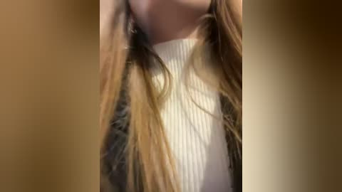 Media: A close-up video of a woman's neck and shoulders, showcasing her long, straight, light brown hair cascading over a white, ribbed turtleneck sweater. The background is a blurry, warm-toned gradient.