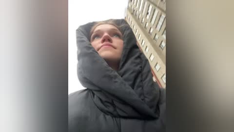 Media: Video of a woman in a black hoodie, arms raised, standing in a tall building's window, looking up. The building's beige facade and many windows are visible.