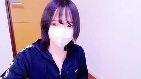 Media: Video of an East Asian woman with straight, shoulder-length dark hair and pale skin, wearing a black jacket, white surgical mask, and seated indoors against a yellow door and white wall.