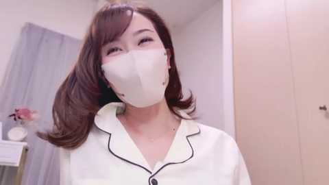 Media: Video of an East Asian woman with long, straight brown hair, wearing a white surgical mask and a white lab coat with black trim. She stands in a softly lit room with white walls, a gray curtain, and a vase of pink flowers.