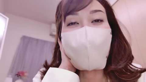 Media: Video of an East Asian woman with long brown hair, wearing a white face mask, touching her chin, in a room with light-colored walls and a grey curtain.