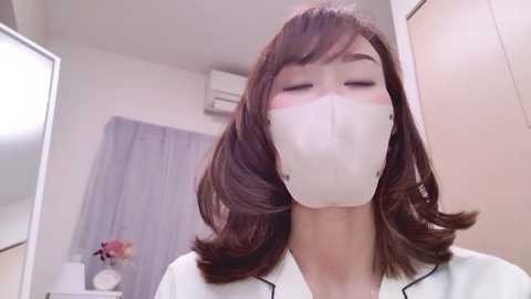 Media: Video of an East Asian woman with shoulder-length brown hair, wearing a white surgical mask and a white coat, in a sterile, brightly lit medical setting.