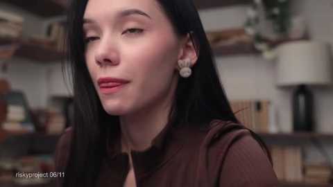 Media: Video of a woman with long black hair, fair skin, and rosy cheeks, wearing a brown top, sitting indoors with a blurred background of shelves and a lamp.