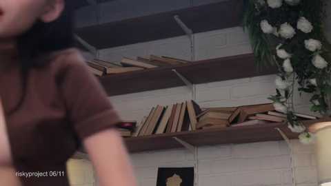 Media: Video of a woman in a brown shirt, standing in a dimly lit room with white brick walls. Shelves hold various books and plants.