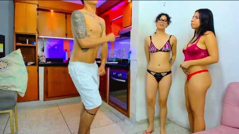 Media: Video of a shirtless man with tattoos, wearing white shorts, dancing in a modern kitchen with two women in lingerie, one in a red top, the other in black.