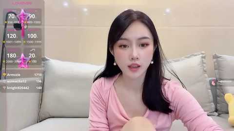 Media: Video of an East Asian woman with long black hair, pink top, and white pearl earrings, seated on a white couch in a minimalist room.