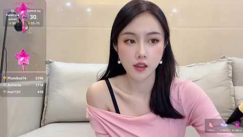 Media: Video of an East Asian woman with straight black hair, wearing a pink off-shoulder top, sitting on a beige sofa. The background includes a pink star overlay and social media icons.