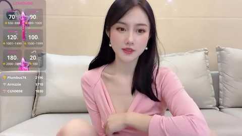 Media: Video of an East Asian woman with long black hair, fair skin, wearing a pink cardigan, sitting on a beige sofa. The background shows virtual health stats overlay.