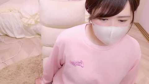 Media: Video of an Asian woman with fair skin, dark hair in a bun, wearing a white face mask, pink long-sleeve shirt with a small logo, sitting on a beige carpet in a softly lit room.