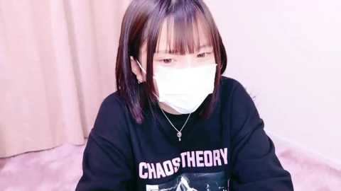 Media: A video of a young Asian woman with straight, shoulder-length black hair and bangs, wearing a black sweatshirt with \"CHAOS THEORY\" in white letters, a white mask, and a silver necklace. She sits on a light pink carpet, against a beige curtain backdrop.