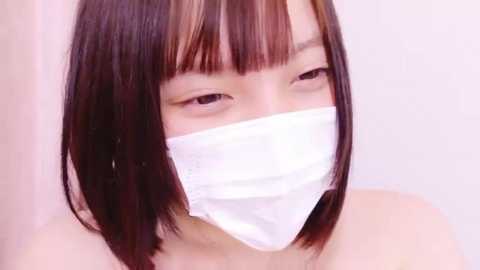 Media: Video of an East Asian woman with straight, shoulder-length dark hair, wearing a white surgical mask, looking downwards, with a plain white background.