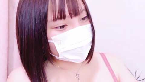 Media: Video of a young Asian woman with straight, shoulder-length dark hair, wearing a white surgical mask and a pink spaghetti-strap top, against a plain white background.