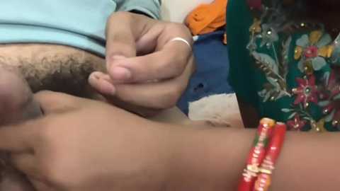 Media: Video of a man's hairy chest with a gold ring on his finger, wearing a light blue shirt. Background includes colorful fabrics and a red bracelet.