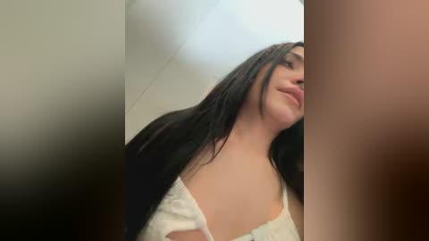 Media: A video of a woman with long black hair, fair skin, and a white blouse, partially cropped from the sides, taken indoors in a dimly lit room.