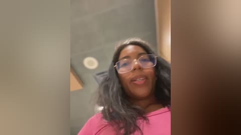 Media: A video of a Black woman with long, wavy black hair, wearing glasses and a pink top, captured indoors. Background features a blurred, beige wall and a light fixture.