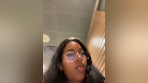 Media: Video of a young Black woman with long, straight black hair and large, clear-framed glasses, wearing a black top, standing indoors near a beige wall with a wooden door and metal railing.