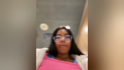 Media: Video of a young Black woman with long straight hair, wearing glasses, a pink shirt, and a white jacket, standing in a bathroom with a beige background.