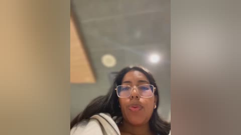 Media: Video of a young woman with medium-dark skin and long black hair, wearing clear glasses and a beige sweater, in a beige hallway with a wooden ceiling.