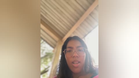 Media: Video of a young woman with medium brown skin, long dark hair, wearing glasses, standing under a wooden-roofed structure with blurred greenery in the background.