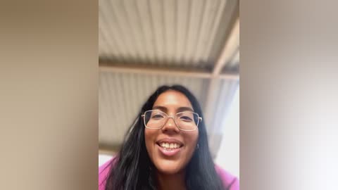 Media: Video of a smiling, light-skinned Black woman with long, straight black hair, wearing clear glasses, and a pink top, indoors with a corrugated metal ceiling.